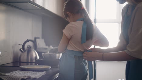 little-girl-is-learning-to-cook-mother-is-putting-apron-on-daughter-child-is-standing-near-stove